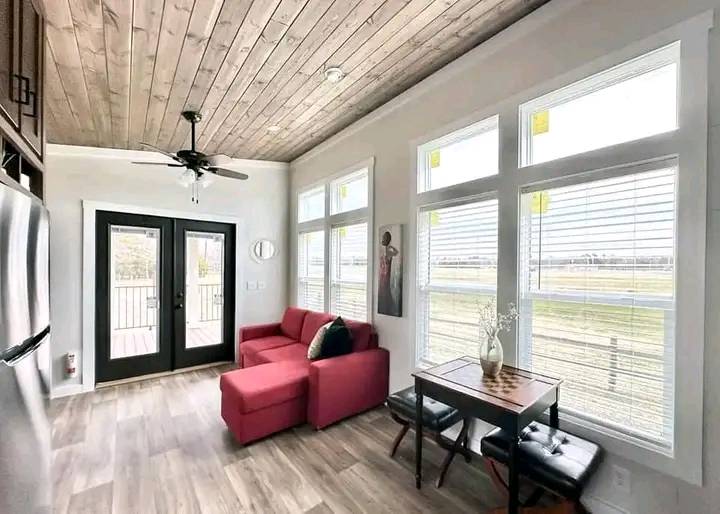 Bright, modern room with a wooden ceiling, fan, and large windows. Features a red sofa, small table with chairs, and black-framed glass doors. Cozy atmosphere.