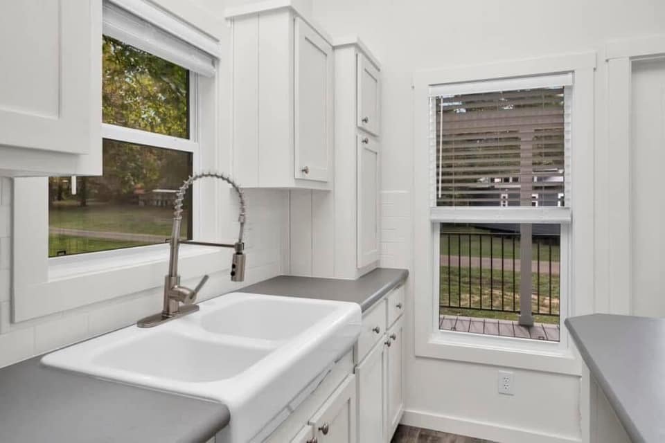 Modern luxury tiny home kitchen featuring pristine white shaker-style cabinetry and a farmhouse-style double sink with a professional-grade gooseneck faucet, showcasing grey quartz-look countertops and large windows that offer a clear view of the covered porch and surrounding nature for a high-end, move-in ready turn-key residential design.