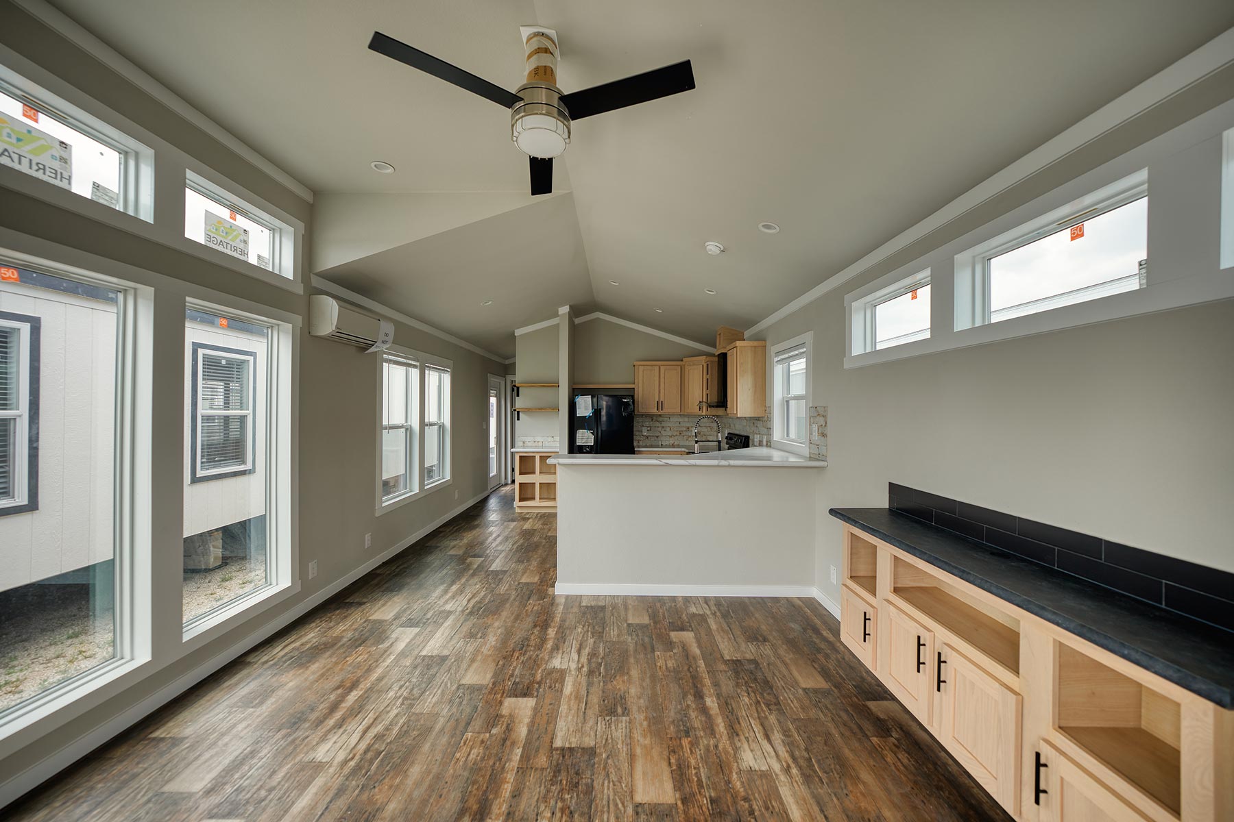 Modern luxury farmhouse-style park model tiny home interior featuring an expansive open-concept living area with premium wide-plank wood-style flooring, high vaulted ceilings, and a contemporary black four-blade ceiling fan. The sophisticated architectural design showcases natural wood cabinetry in the gourmet kitchen, elegant white marble-patterned countertops with a breakfast bar, and sleek black hardware throughout the minimalist space. Large energy-efficient windows and transom lights flood the professional "full rank" SEO-optimized layout with natural light, highlighting the built-in media console with a black soapstone-style top and high-end stainless steel appliances. This high-end prefab residence offers a perfect showcase for custom-built modular homes, luxury vacation cottages, or modern backyard accessory dwelling units (ADU) designed for maximum style, functional elegance, and a bright, airy aesthetic.