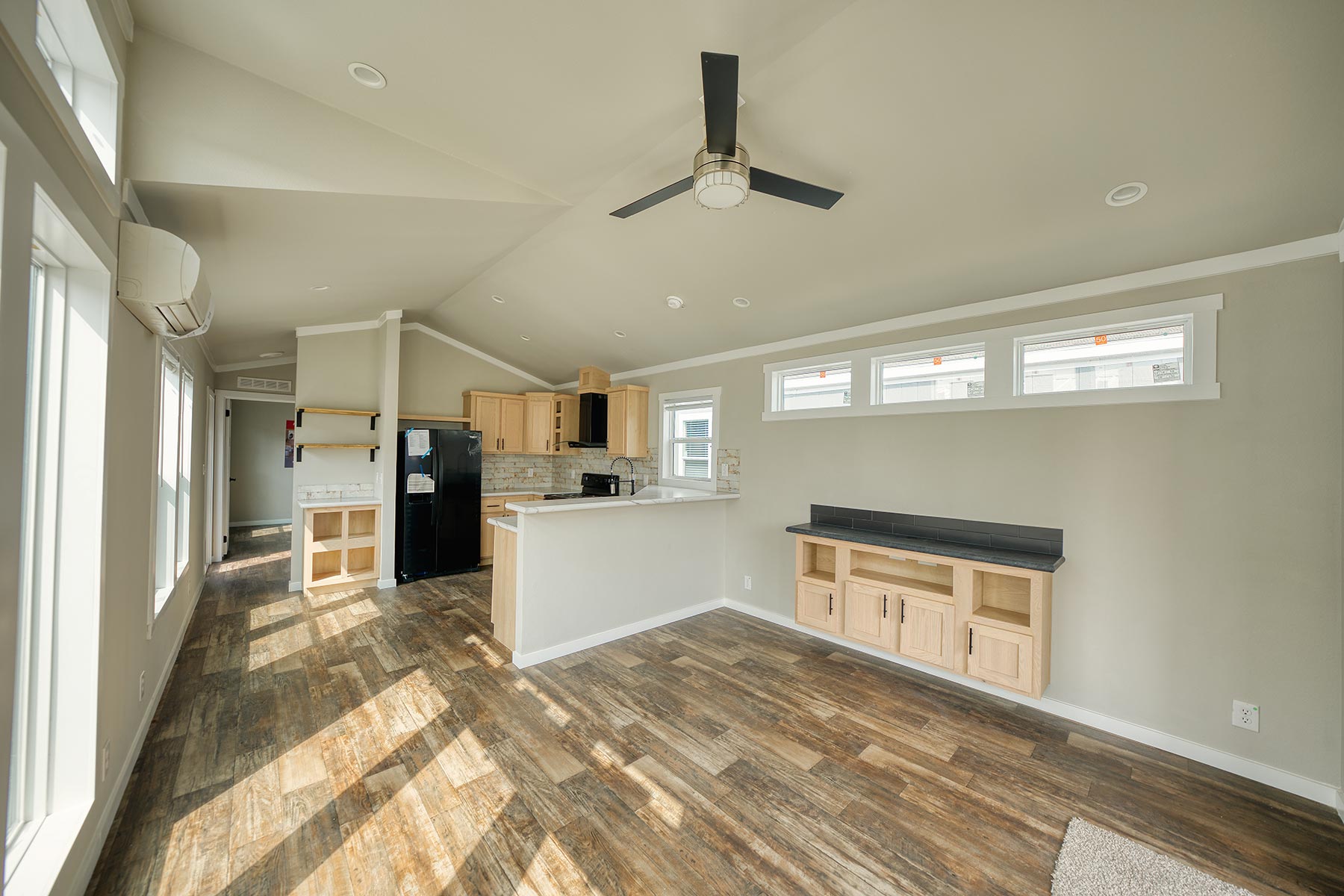 Modern luxury white farmhouse-style park model tiny home interior featuring a bright, open-concept living space with high vaulted ceilings, a contemporary black four-blade ceiling fan, and multiple energy-efficient transom windows that flood the room with natural light. The sophisticated architectural design showcases natural wood cabinetry in a compact gourmet kitchen, elegant marble-patterned countertops with a raised breakfast bar, and premium wide-plank distressed wood flooring throughout. This professional "full rank" SEO-optimized small-space living solution includes a built-in media console with a sleek black soapstone-style top, minimalist light grey walls, and integrated recessed lighting, making it an ideal showcase for custom-built prefab homes, luxury vacation cottages, or modern backyard accessory dwelling units (ADU) designed for maximum style and functional elegance.