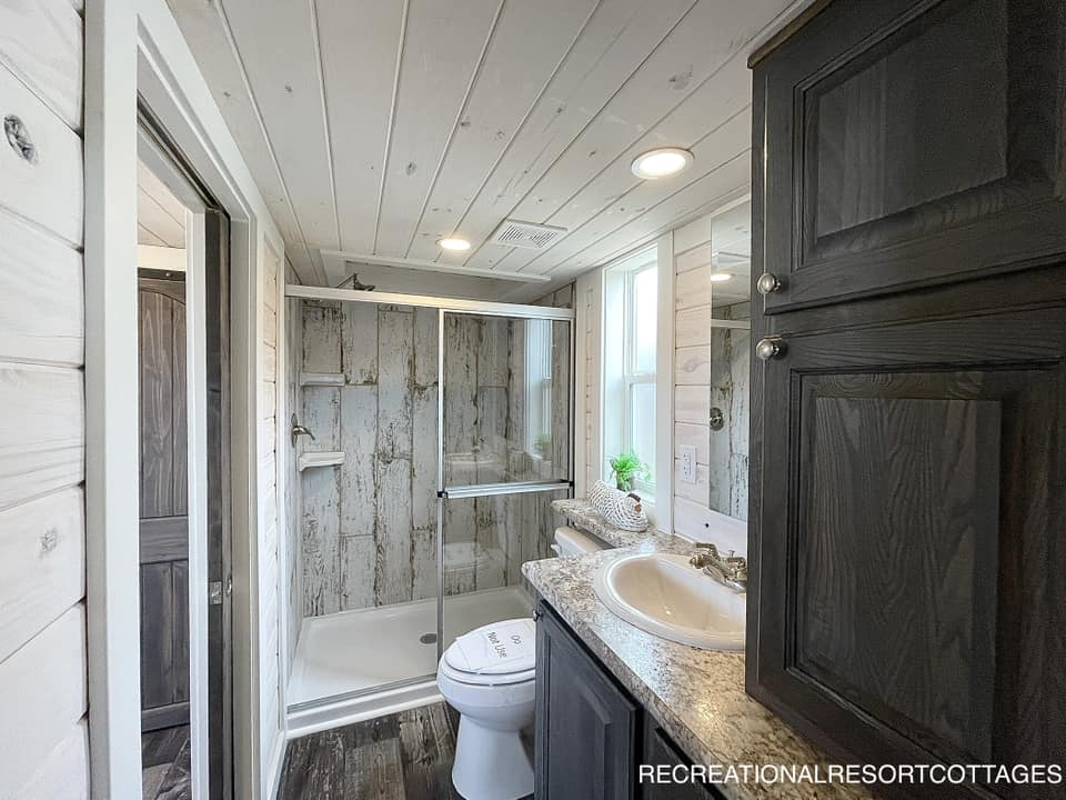 Compact modern farmhouse bathroom with wood walls, a stone countertop, and dark cabinets. A glass shower with barnwood-style panels and a small sink area enhance coziness.