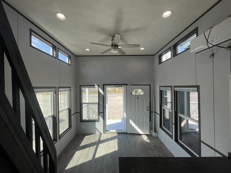 Modern tiny home interior, featuring large windows, high ceiling with recessed lighting, ceiling fan, and a white front door. Bright and airy ambiance.