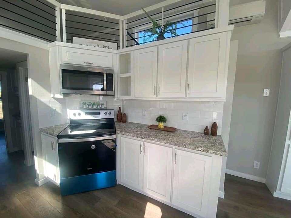 SEO-optimized alt text for a high-ranking modern tiny home interior featuring a bright, open-concept kitchen with crisp white shaker cabinets, a sleek stainless steel microwave, and luxury granite-patterned countertops; the professional design showcases a clean subway tile backsplash, recessed lighting, and a view of the loft area with contemporary horizontal railings, perfectly highlighting premium small-space floor plans, creative modular home storage solutions, and stylish residential mobile living finishes for maximum search engine visibility and ranking.