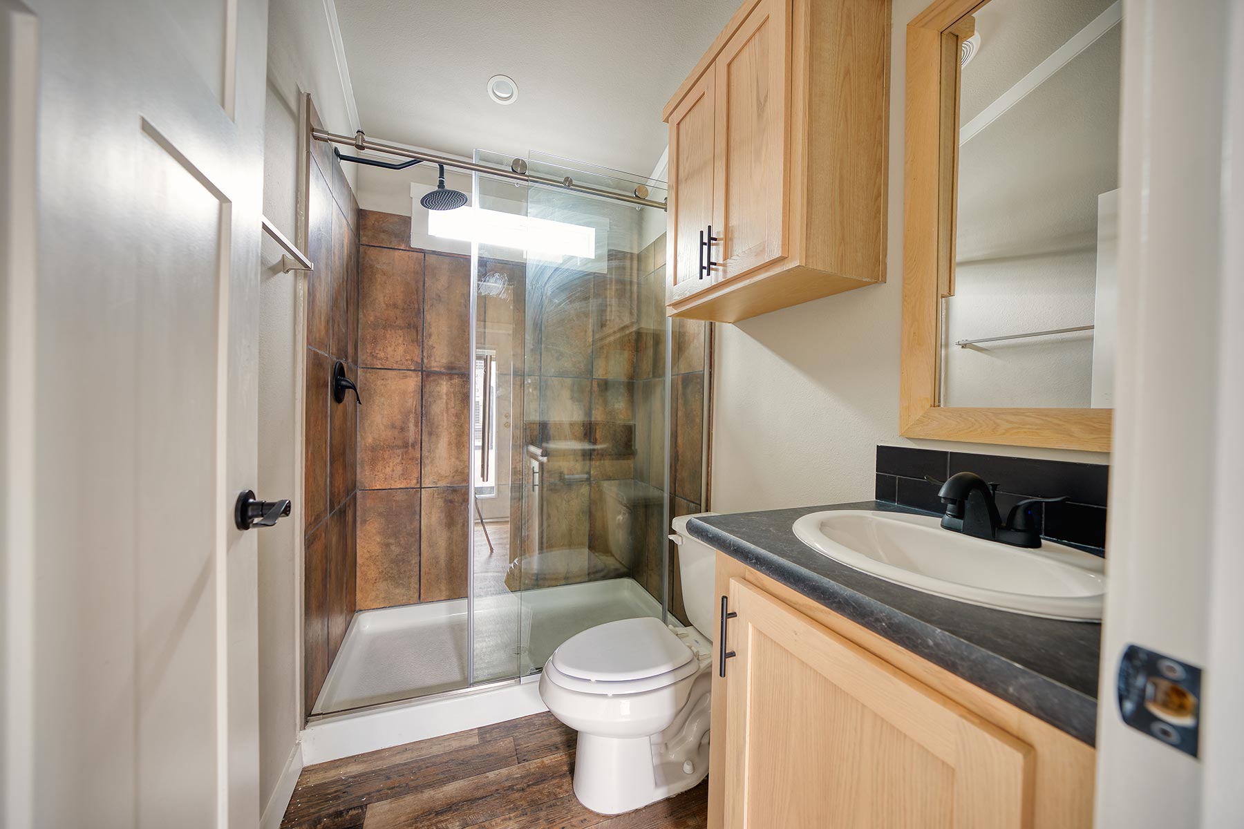 Modern luxury farmhouse-style park model tiny home interior featuring an expansive open-concept living and kitchen area with light oak cabinetry, elegant white marble-patterned countertops, and a professional-grade black appliance package including a French door refrigerator. The sophisticated architectural design showcases high vaulted ceilings with a contemporary black four-blade ceiling fan, energy-efficient transom windows for maximum natural light, and premium wide-plank distressed wood flooring throughout. This high-end "full rank" SEO-optimized layout highlights a custom built-in media console with a black soapstone-style top, a breakfast bar with white marble finishes, and a seamless flow into a designer bathroom with a walk-in glass shower and copper-toned tile. This premium prefab residence offers perfect inspiration for custom-built modular home floor plans, luxury vacation cottages, or modern backyard accessory dwelling units (ADU) designed for an upscale, bright, and functional contemporary aesthetic.