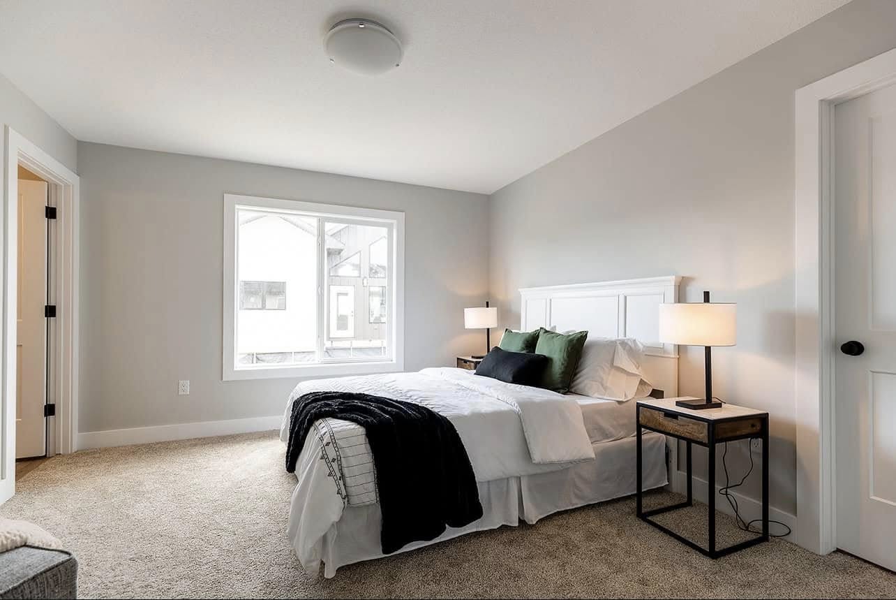 Modern bedroom with a cozy, neutral tone. Features a white bed, green and black cushions, bedside tables with lamps, carpet flooring, large window, and soft lighting.