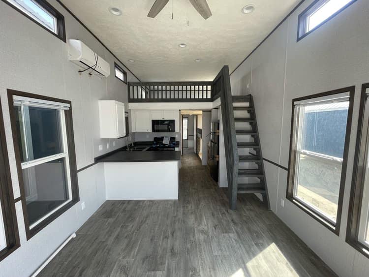 Spacious tiny home interior with light wood flooring, loft space, sleek modern kitchen, gray staircase, large windows, and neutral tones.