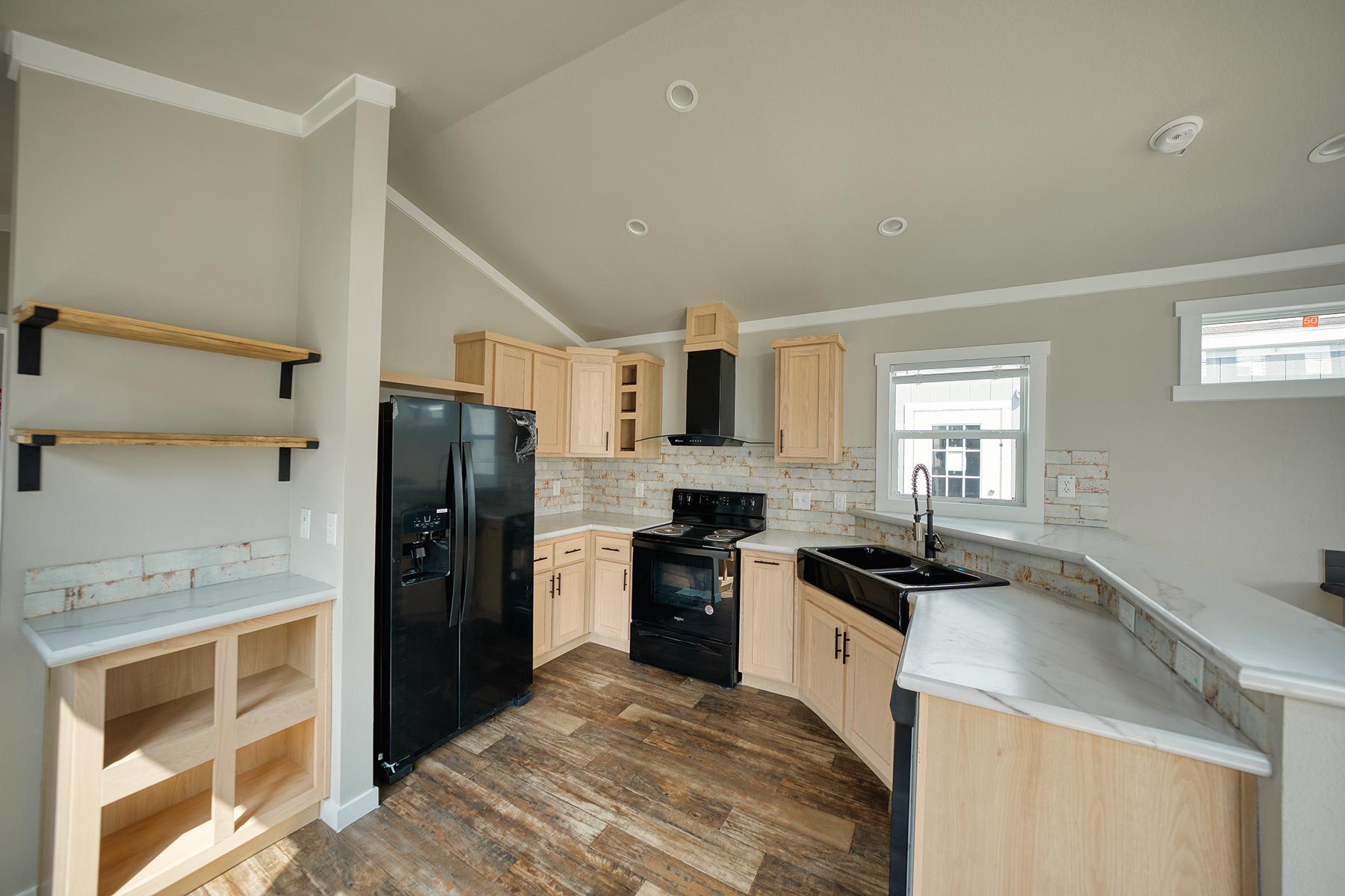 SEO-optimized alt text for a modern luxury manufactured home or park model interior, featuring an open-concept living area with vaulted ceilings, a sleek black ceiling fan, and rustic wood-look vinyl flooring; the space showcases a gourmet kitchen with natural wood-tone cabinetry, marble-style countertops, and high-end black appliances, complemented by expansive windows and transom windows that flood the room with natural light; this premium small-space design highlights contemporary architectural details such as a mini-split HVAC system, neutral gray walls, and innovative storage solutions, perfectly suited for high-ranking searches related to luxury mobile living, modular home floor plans, and stylish tiny house interior decor.