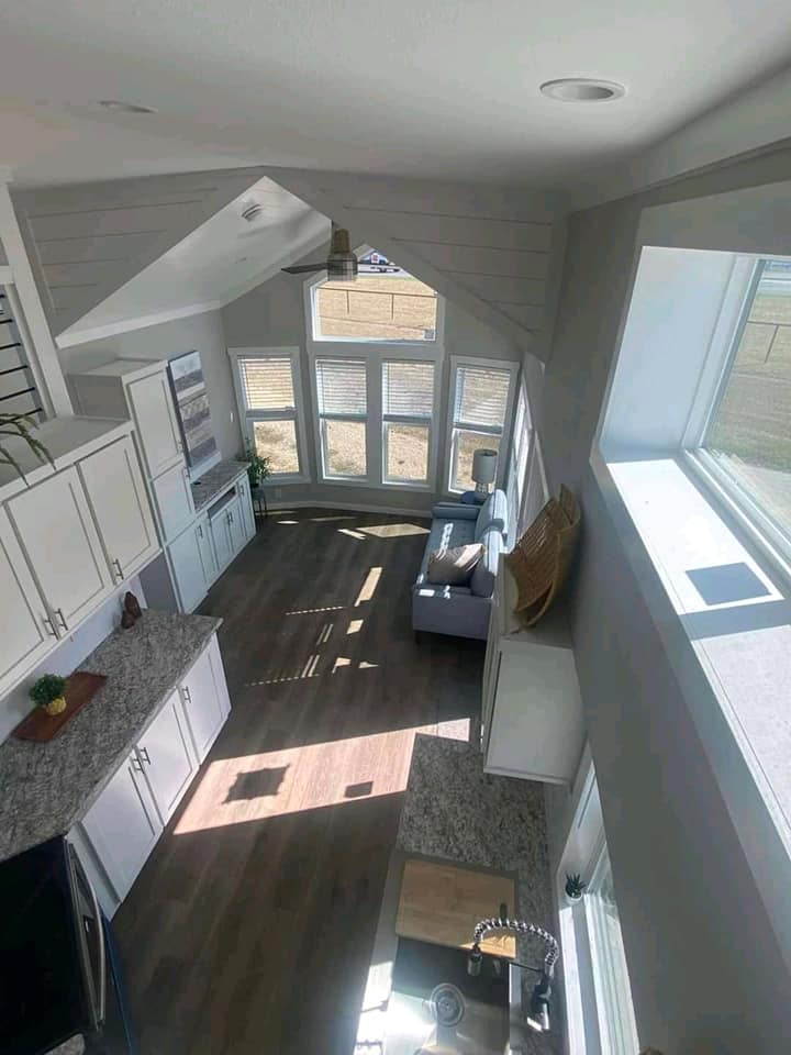 SEO-optimized alt text for a high-ranking modern tiny home or luxury park model interior featuring an aerial loft view of a bright, open-concept living area with vaulted white shiplap ceilings and recessed lighting; the professional interior design showcases a contemporary galley kitchen with white shaker cabinets and granite countertops, a cozy blue sofa nestled under large panoramic windows, and durable dark wood-plank flooring, perfectly highlighting premium small-space floor plans, creative modular home layouts, and stylish residential mobile living solutions for maximum search engine visibility and ranking.