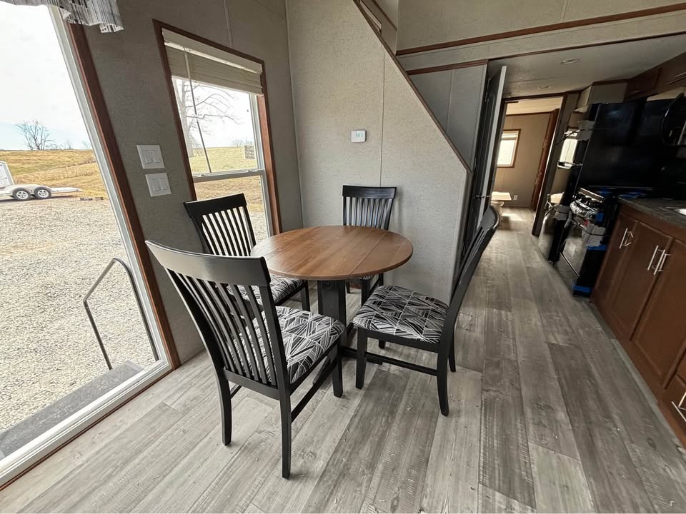 Cozy kitchen nook with a round wooden table and four patterned chairs, set on light wood flooring. A staircase and modern kitchen are visible in the background. Natural light streams in through a large window, creating a warm, inviting atmosphere.