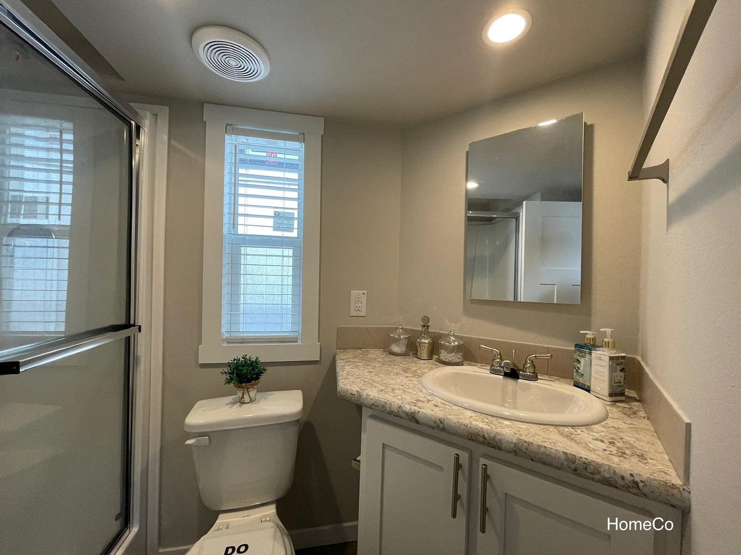Compact bathroom featuring a glass-enclosed shower, a white toilet with a small potted plant, a vanity with a speckled countertop and white cabinets, a stainless steel faucet, and a mirror above the sink, all under a recessed light and ceiling vent.