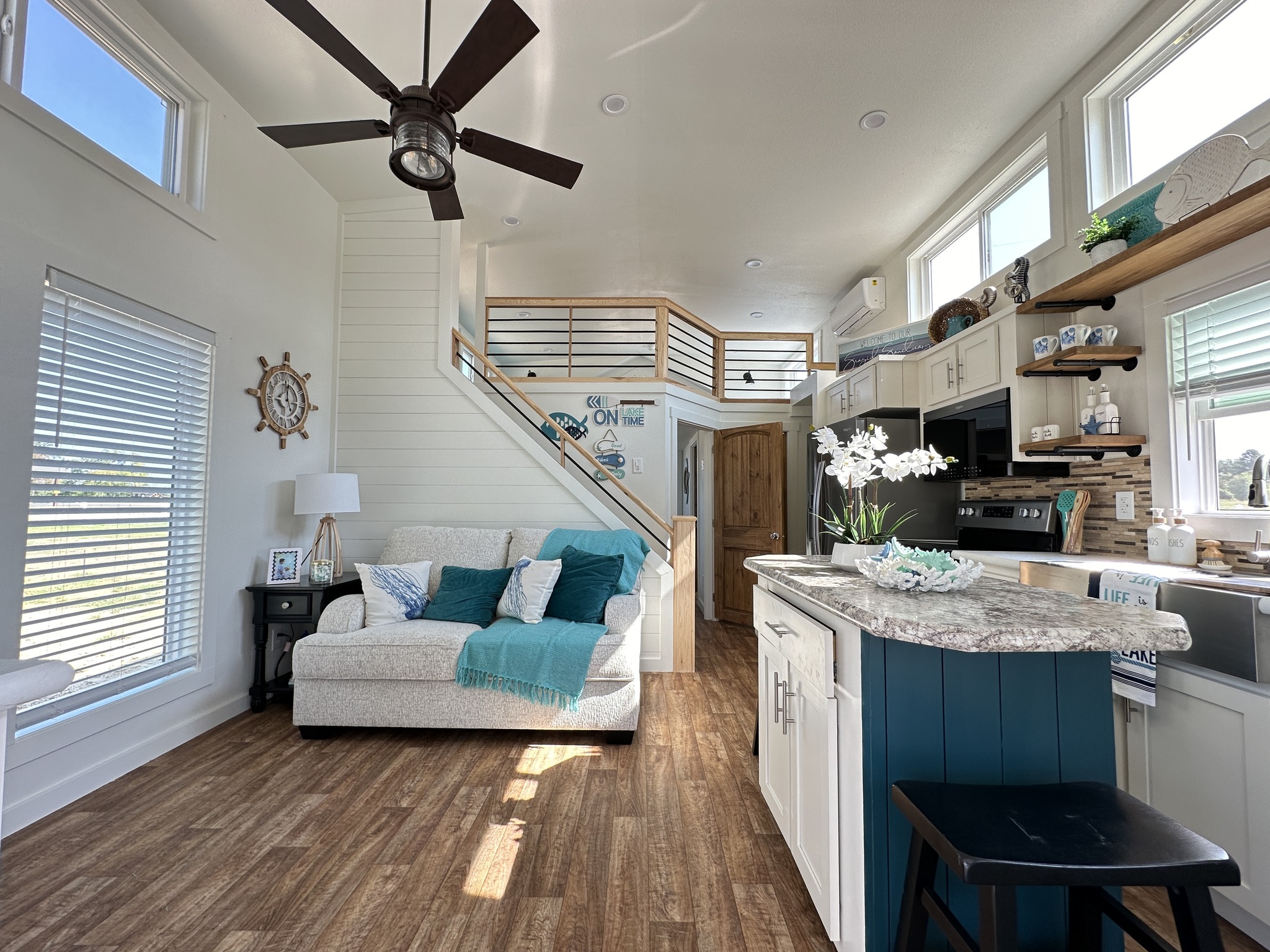 Modern luxury nautical-themed tiny home interior featuring a bright, open-concept living area with a high-end vaulted ceiling, clerestory windows, and a contemporary dark wood ceiling fan. The sophisticated space showcases a cozy white sofa with teal accents, a sleek loft area with industrial wire railings, and a gourmet kitchen with a vibrant teal island, marble-style countertops, and stainless steel appliances, all set on premium wide-plank flooring to create a perfect "full rank" SEO-optimized aesthetic for luxury prefab or custom-built small-space living.