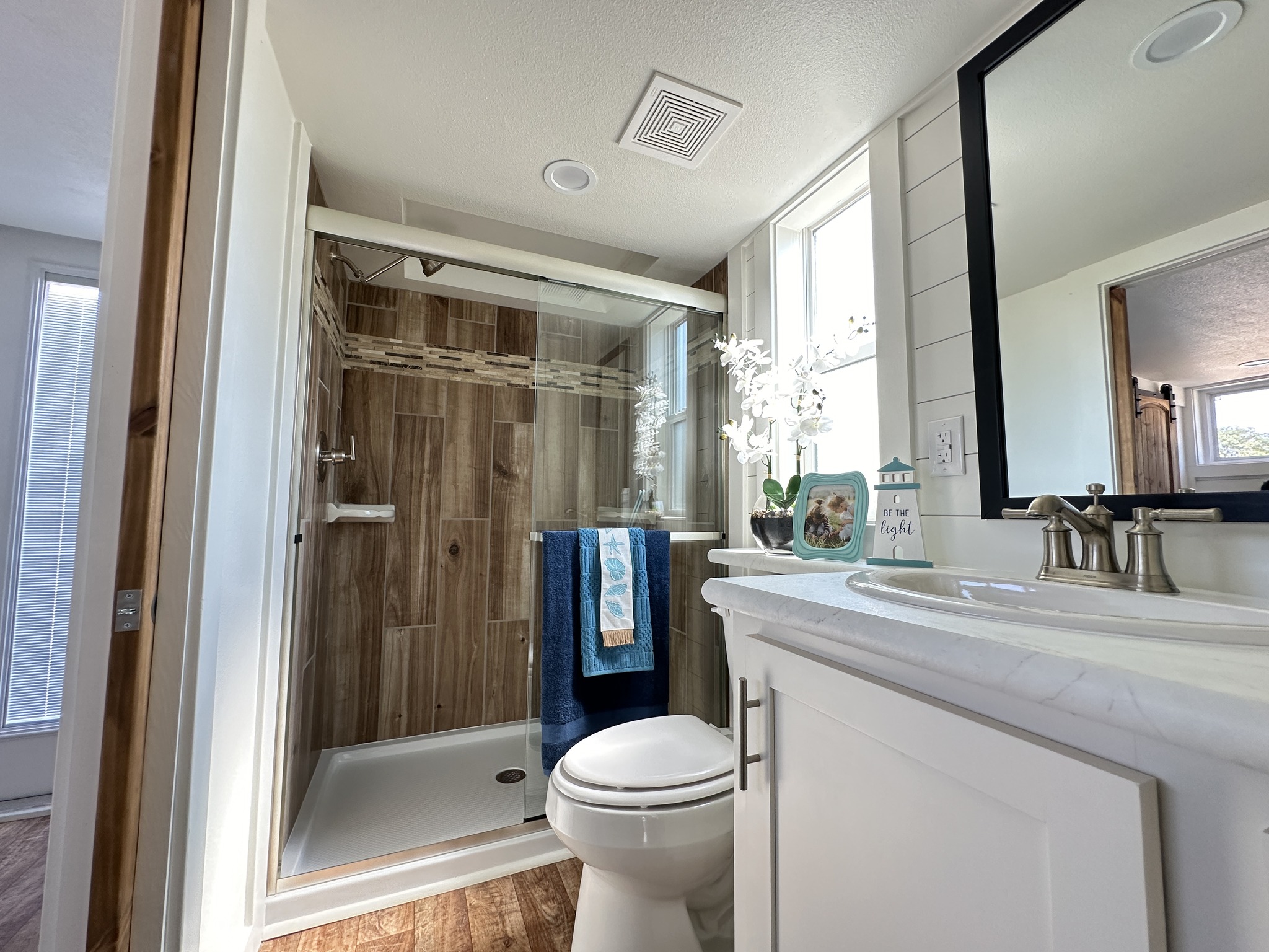 Modern luxury tiny home bathroom interior featuring a bright and airy coastal theme, showcasing a spacious walk-in shower with vertical wood-look tile surrounds and a sliding glass door, alongside a sleek white vanity with a marble-patterned countertop and a large black-framed mirror for a high-end, move-in ready turn-key residential design.