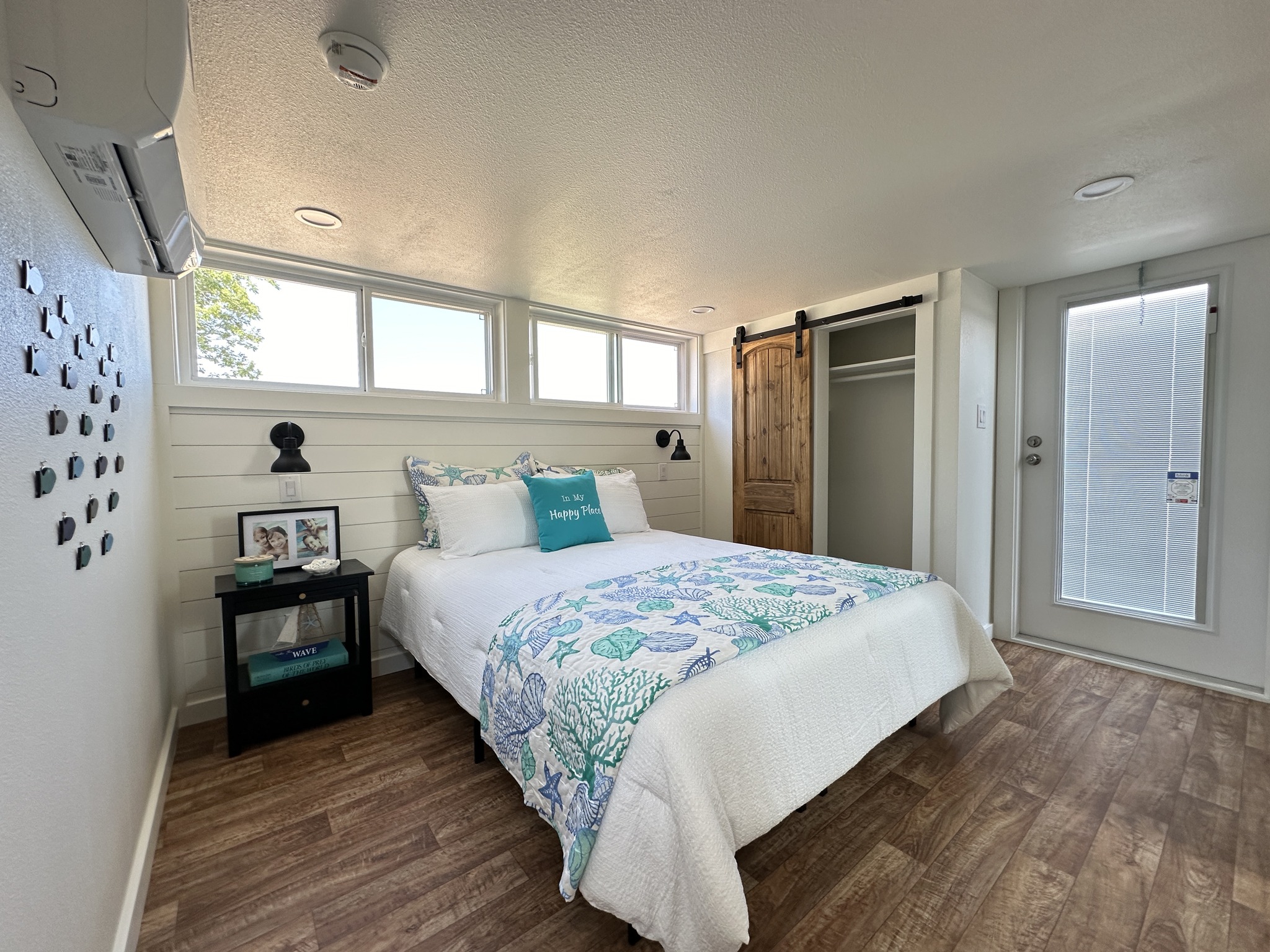 Modern luxury tiny home bedroom with a coastal theme featuring a plush queen-sized bed adorned with teal and white sea-inspired bedding and a "Happy Place" accent pillow. The sophisticated small-space design showcases bright white shiplap walls, a rustic sliding barn door for the closet, and premium wide-plank wood flooring, all illuminated by a row of energy-efficient clerestory windows and contemporary black wall sconces. This high-end interior captures a serene "full rank" SEO-optimized aesthetic, perfect for a luxury prefab vacation cottage, custom-built park model home, or a stylish accessory dwelling unit (ADU) with a professional and airy feel.