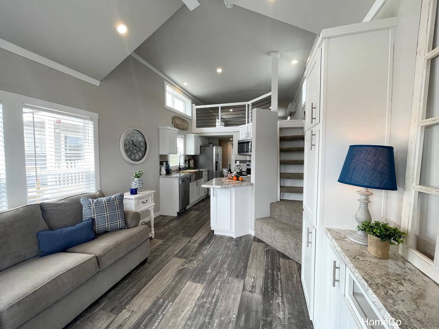 Modern luxury tiny home interior featuring a bright open-concept living area with a grey sofa and blue accent pillows, showcasing a functional white shaker-style kitchen with granite countertops and a space-saving carpeted staircase leading to a private sleeping loft with cable safety railings under a vaulted white shiplap ceiling for a high-end move-in ready turn-key residential design.