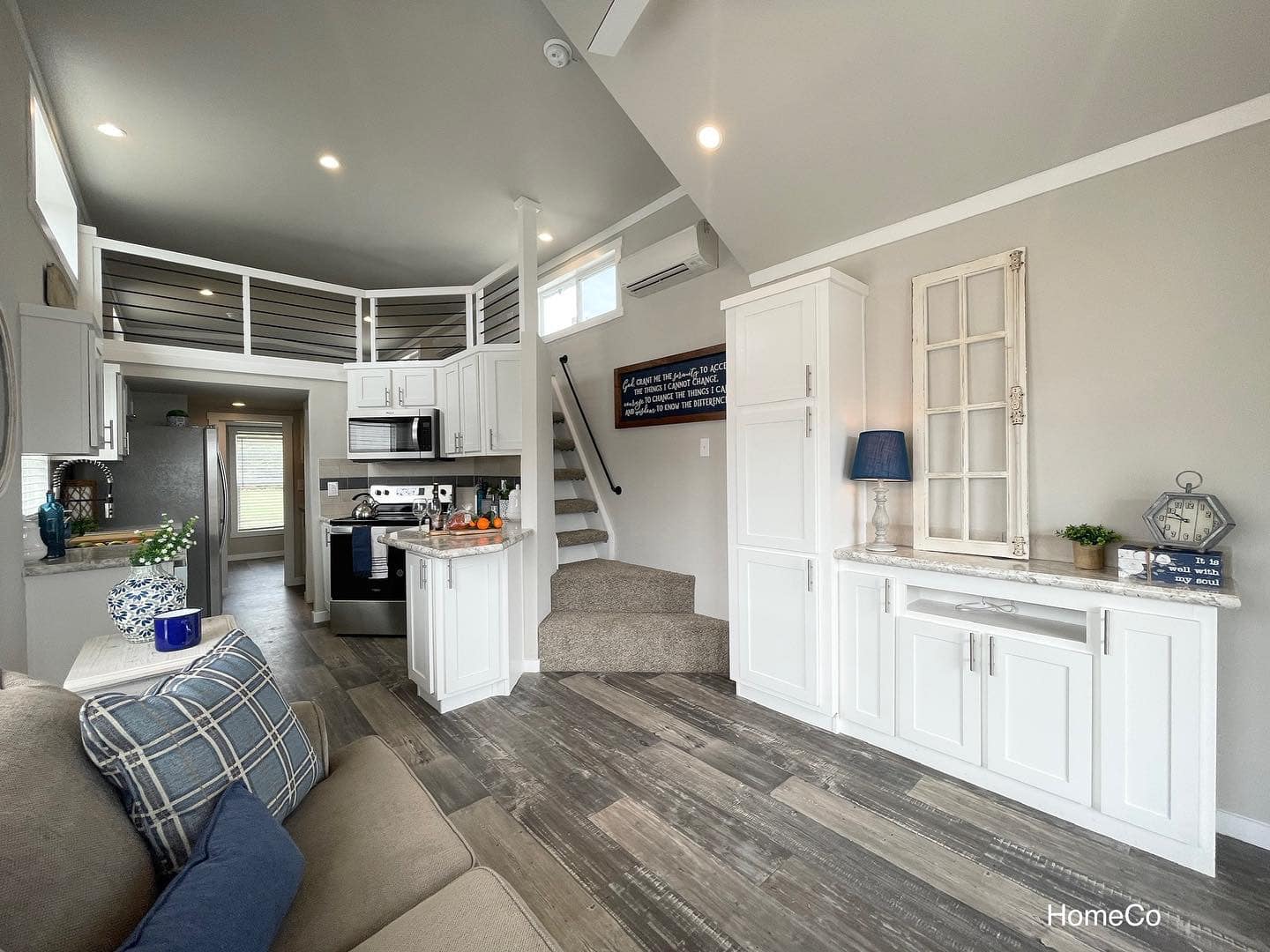 Compact modern living space with gray walls and wood flooring. Features include a kitchen with white cabinets, loft area, and cozy seating. Bright and inviting.