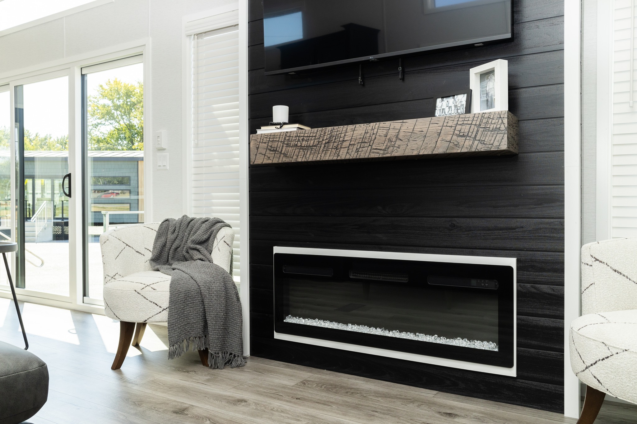 Modern living room with a black feature wall, mounted TV, and electric fireplace. A patterned chair draped with a gray throw sits by a glass sliding door.