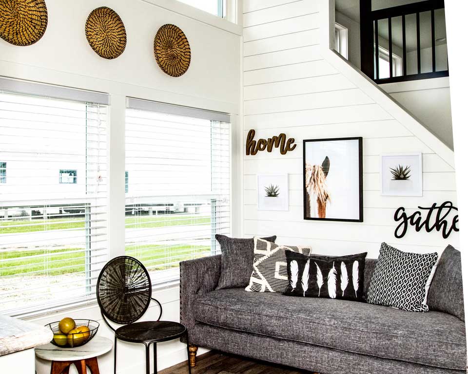 Cozy living room with a grey couch adorned with patterned cushions, white shiplap walls, decorative wall art, and basket decor. Large windows add natural light.