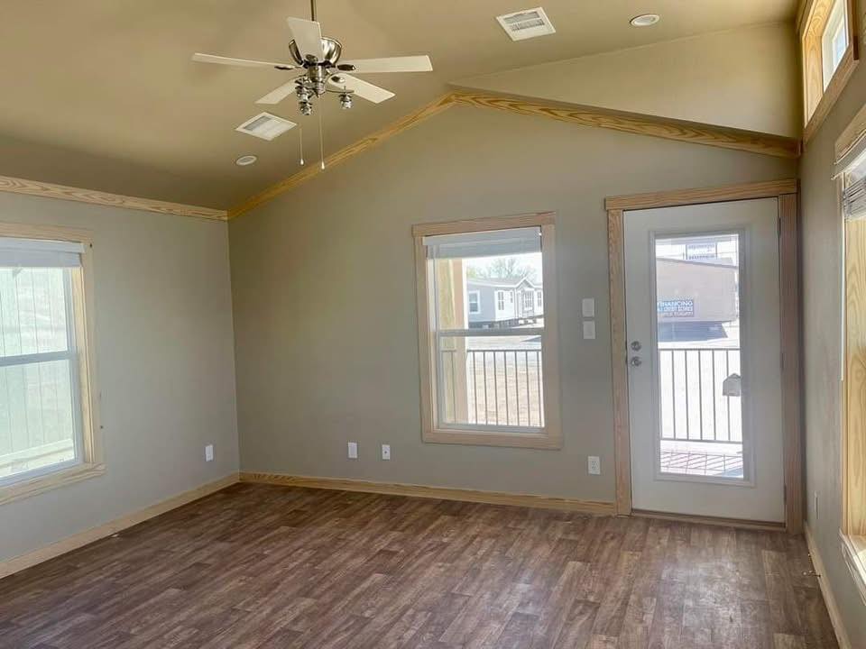 Spacious empty modern farmhouse bedroom in tiny home or modular cottage featuring vaulted tray ceiling with exposed wooden trim accents, ceiling fan with light kit, neutral taupe gray walls, warm medium-tone luxury vinyl plank flooring, large double-hung windows with transom clerestory glass flooding natural light, French patio door with glass panels opening to balcony railing view, white baseboards and door trim, recessed lighting, minimalist airy design ideal for primary suite, guest room, vacation cottage retreat, affordable tiny house living, or customizable modular home interiors.