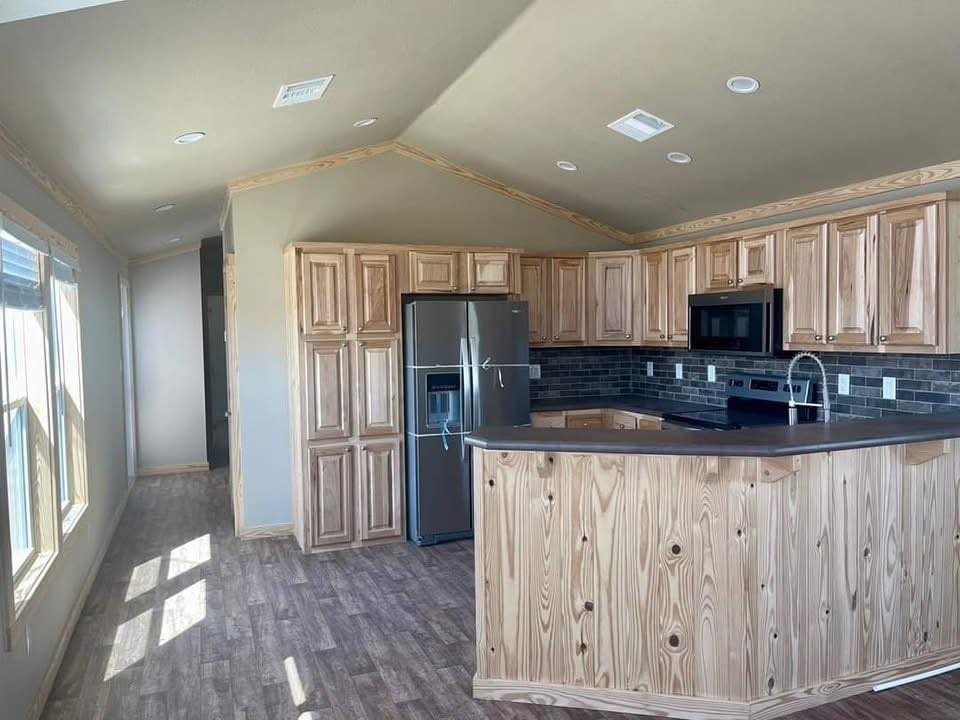 Spacious open-concept modern farmhouse kitchen in a tiny home or modular cottage featuring vaulted ceiling with warm wooden trim accents and recessed lighting, light gray walls, medium-tone luxury vinyl plank flooring, rustic light pine shaker cabinets with natural wood grain finish, large curved breakfast bar island with dark countertop, stainless steel side-by-side refrigerator with water dispenser, black subway tile backsplash, stainless steel gas range and microwave, farmhouse sink with chrome pull-down faucet, abundant natural light from oversized windows overlooking outdoor views, airy bright layout ideal for family gatherings, cozy tiny house living, affordable modular homes, or vacation cottage retreats.