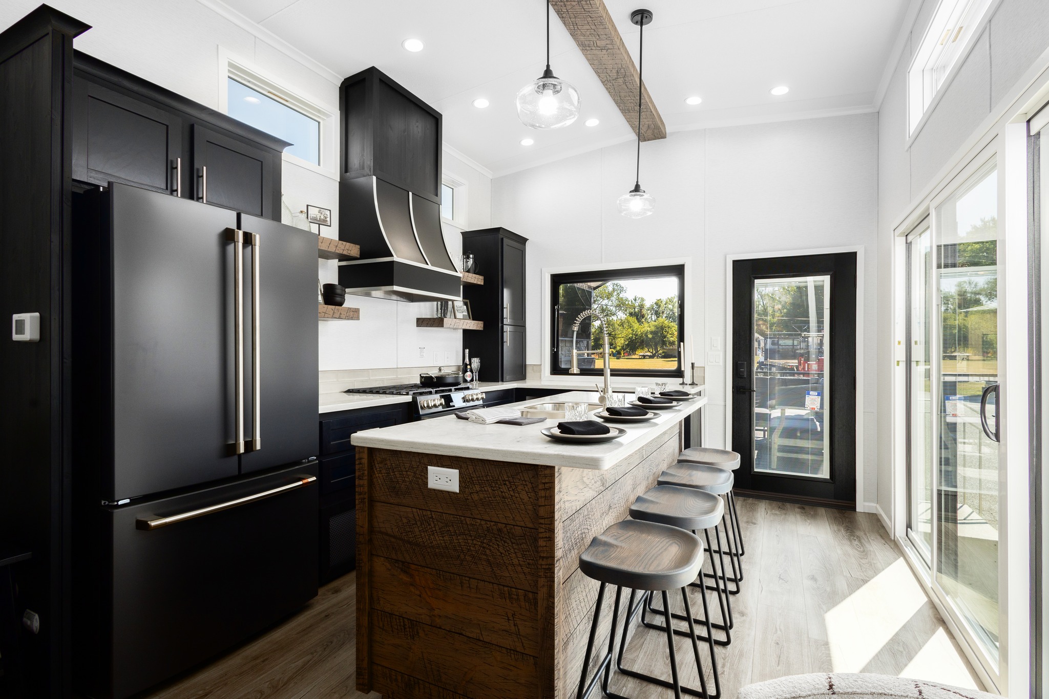 Modern kitchen with a large black fridge, wooden island with bar stools, and sleek fixtures. Sunlight streams through glass doors, creating a bright, inviting atmosphere.