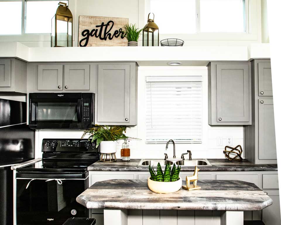 Modern kitchen with gray cabinets, a black stove and microwave, and a rustic island. Decor includes plants, lanterns, and a "gather" sign, creating a cozy vibe.