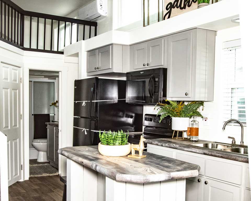 Compact kitchen with gray cabinets, black appliances, and wood-accented island. Green plants add warmth. A bathroom is visible in the background.