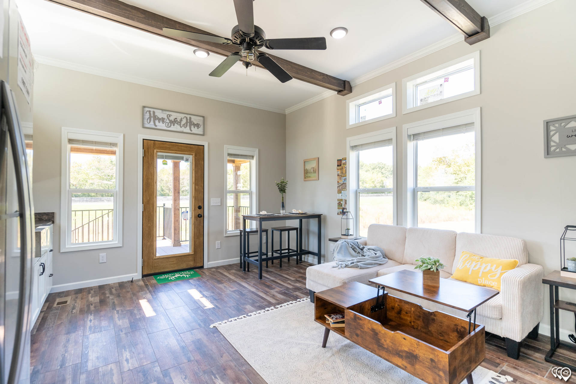 Modern luxury tiny home interior featuring a bright open-concept living area with premium wood-look flooring and rustic dark wood ceiling beams, showcasing a comfortable white sectional sofa, a multifunctional wooden lift-top coffee table, and a dedicated dining nook with a high-top table and stools near a large glass entry door for a sophisticated move-in ready turn-key residential design.