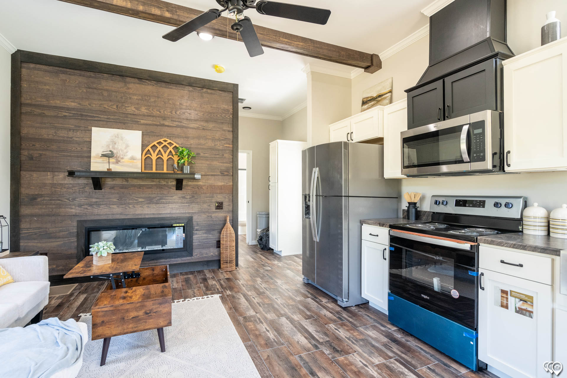 Modern luxury tiny home interior featuring a spacious open-concept living area with a dark wood-plank accent wall and premium wood-look tile flooring, showcasing a contemporary white shaker-style kitchen with a central dark wood island and integrated wine storage under a tray ceiling with a stylish fan for a 100% move-in ready turn-key residential design.