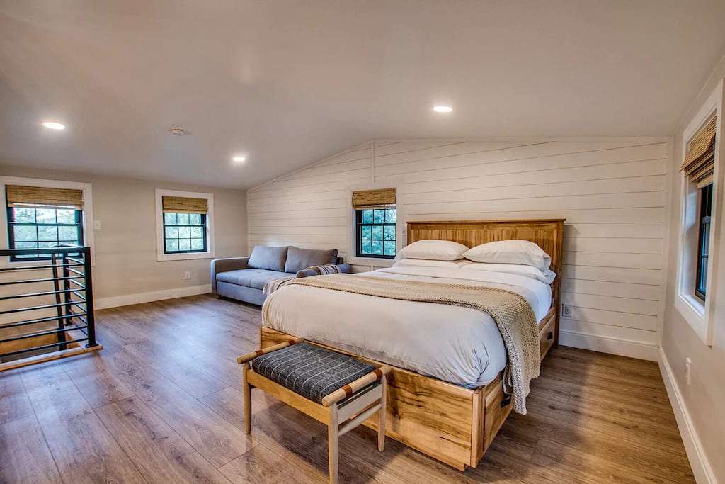 Spacious bedroom with wooden flooring features a cozy bed, soft grey couch, and large windows. Warm, calming ambiance with minimal decor.