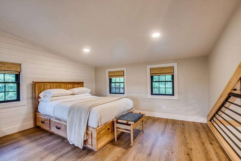 A cozy bedroom with a wooden bed, white bedding, and a beige throw. Three windows with bamboo blinds let in natural light. The room has wooden floors and neutral walls, creating a warm, inviting atmosphere.