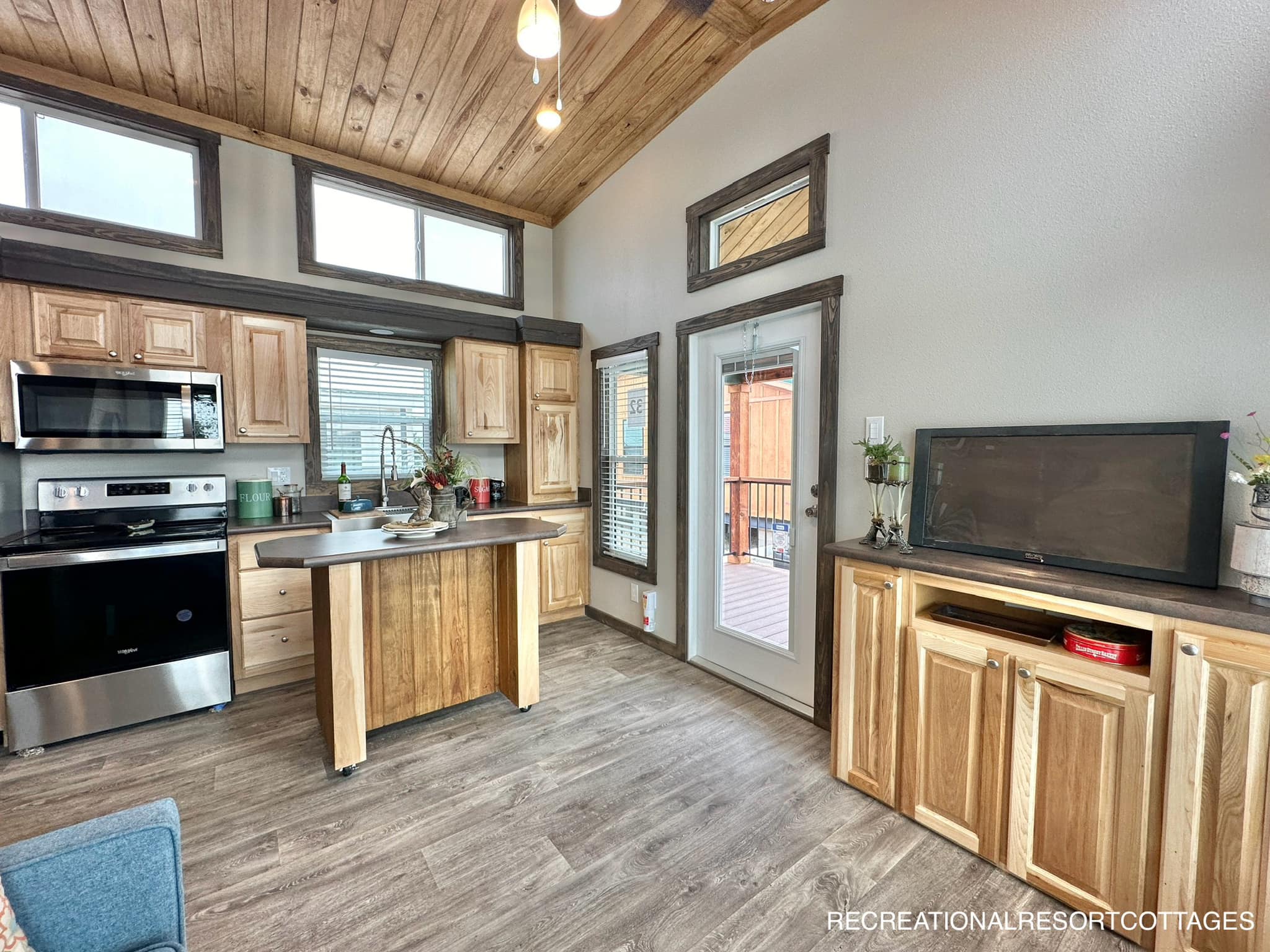 Cozy kitchen with wood cabinets, stainless steel appliances, and a small island. Natural light from high windows adds warmth. Rustic, inviting ambiance.