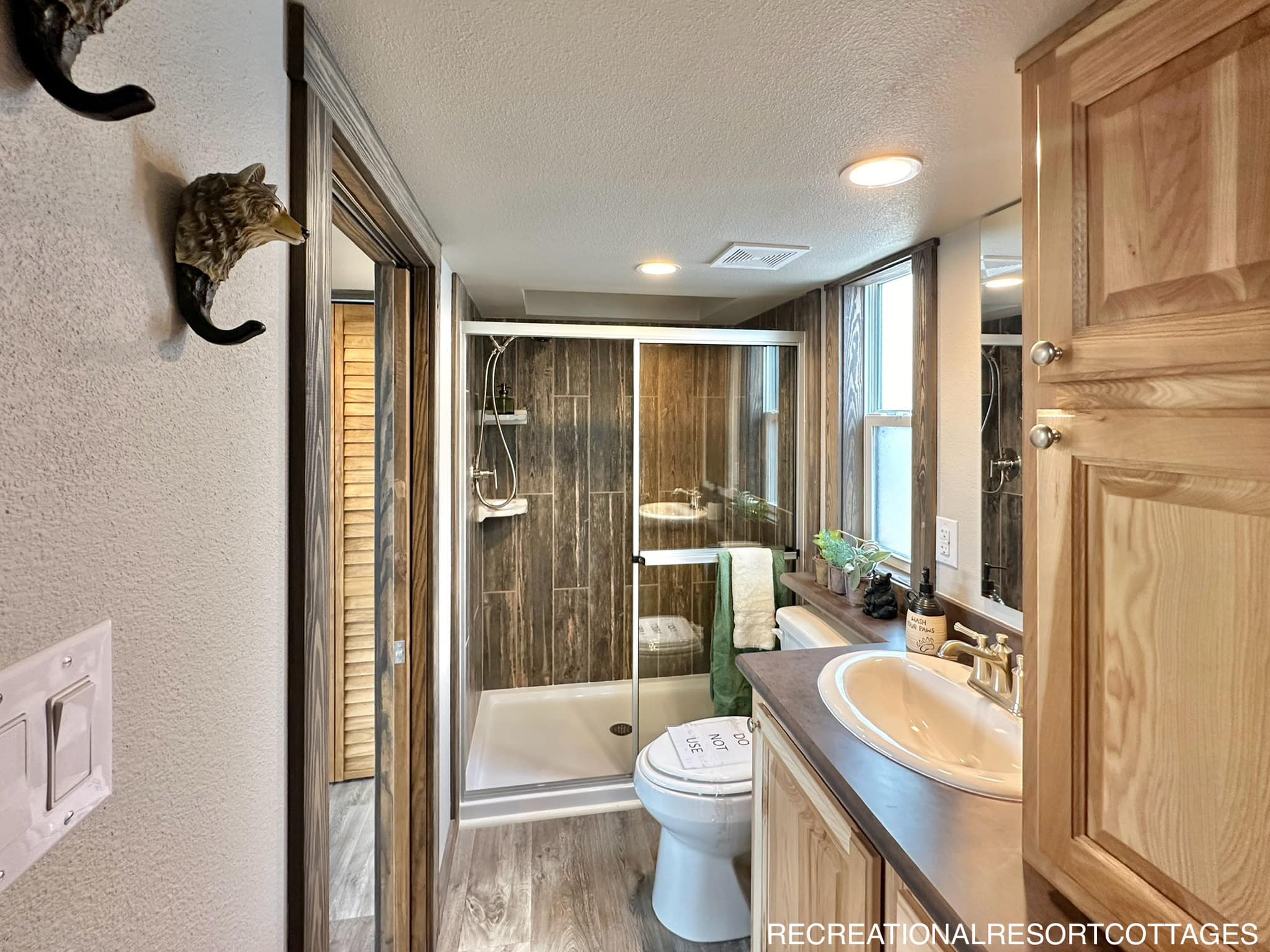 A cozy bathroom with wood accents features a glass-enclosed shower, wooden cabinets, and a sink with a mirror. A toilet is next to a window with natural light. A green towel adds a pop of color.