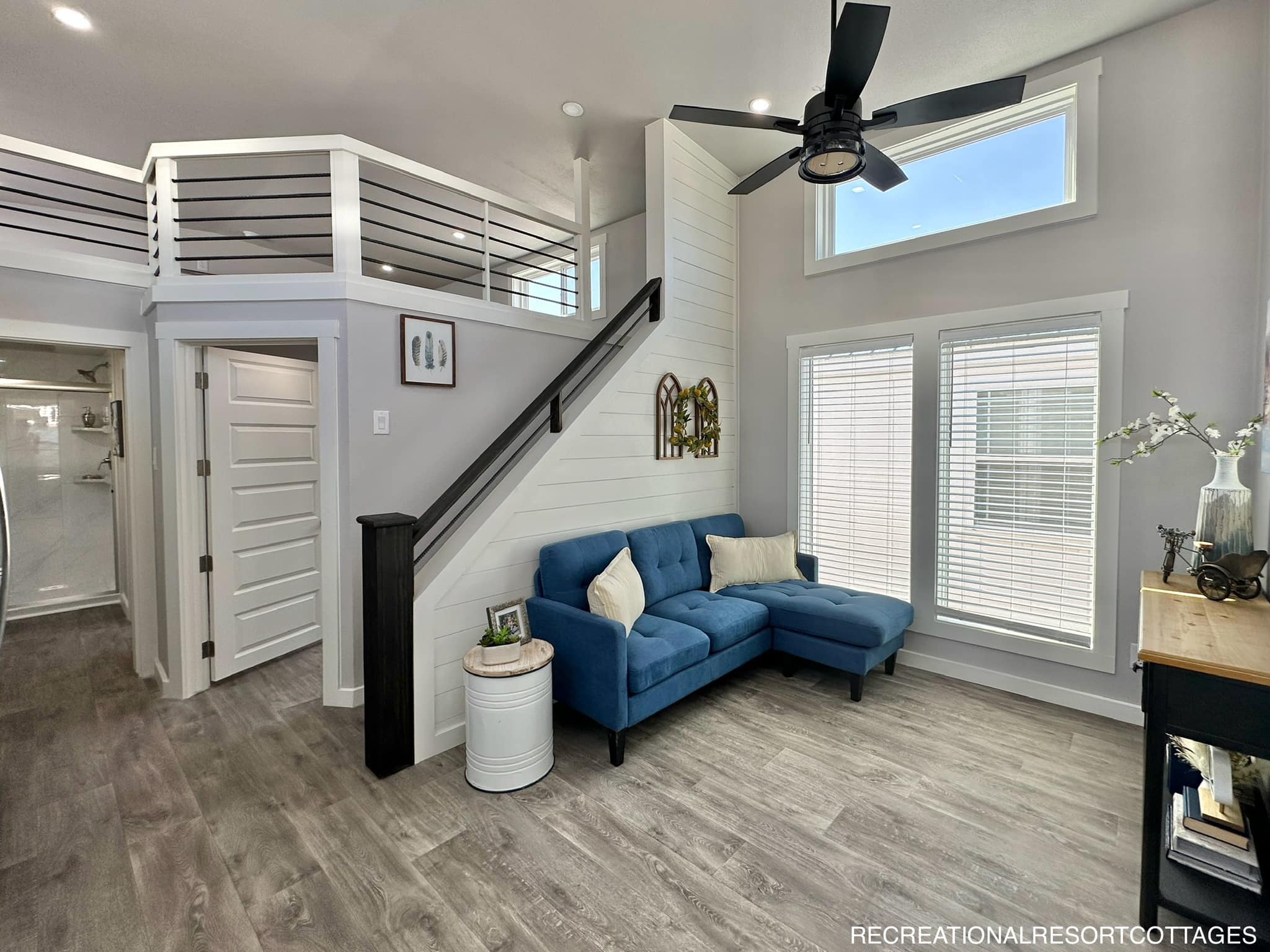 Modern living room with high ceilings, large windows, and a blue sectional sofa. A staircase leads to a loft. The space is bright and airy.