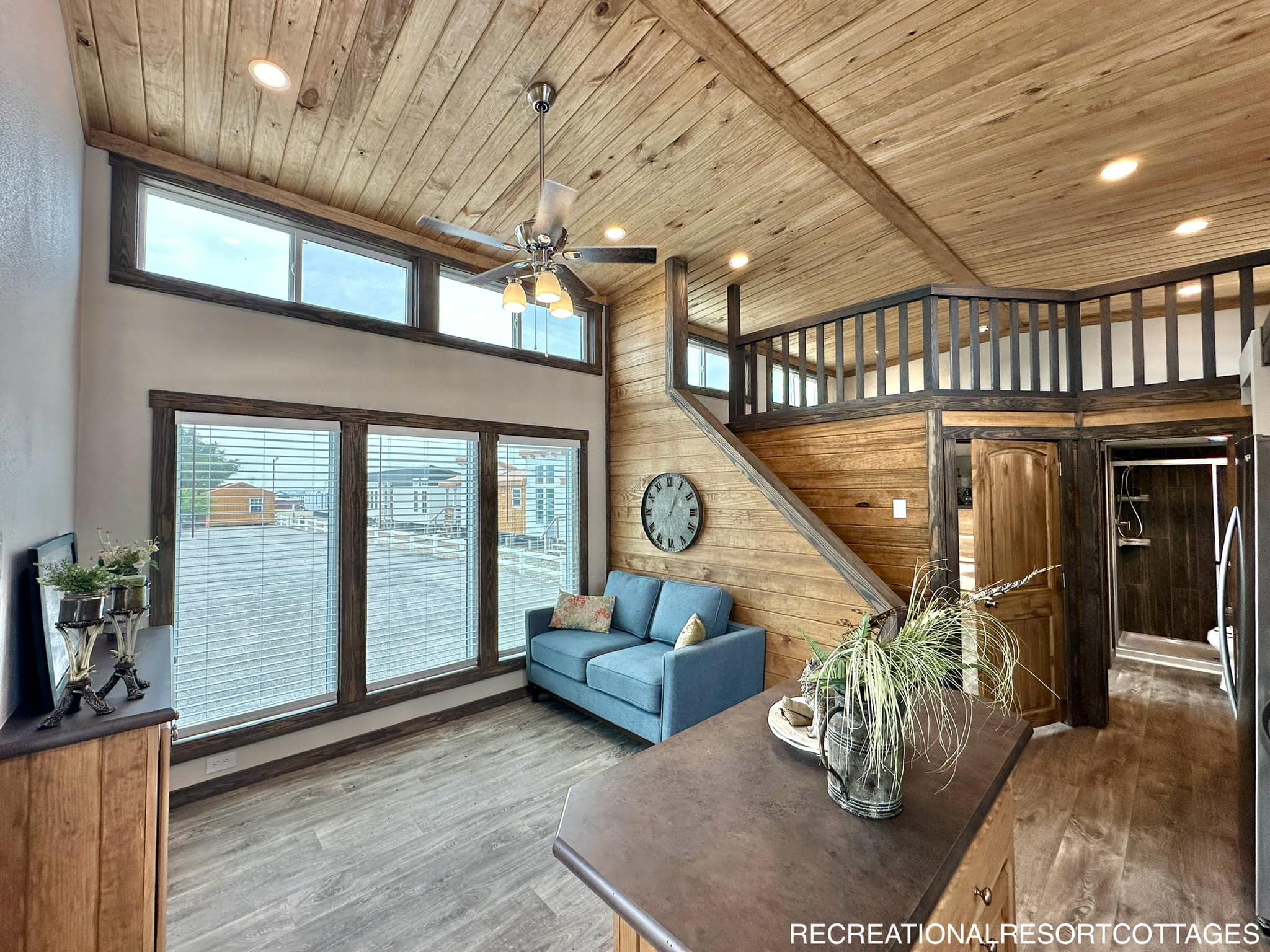 Cozy wooden interior of a cabin with large windows, a blue sofa, and a rustic clock on the wall. Stairs lead to a loft. Warm and inviting atmosphere.