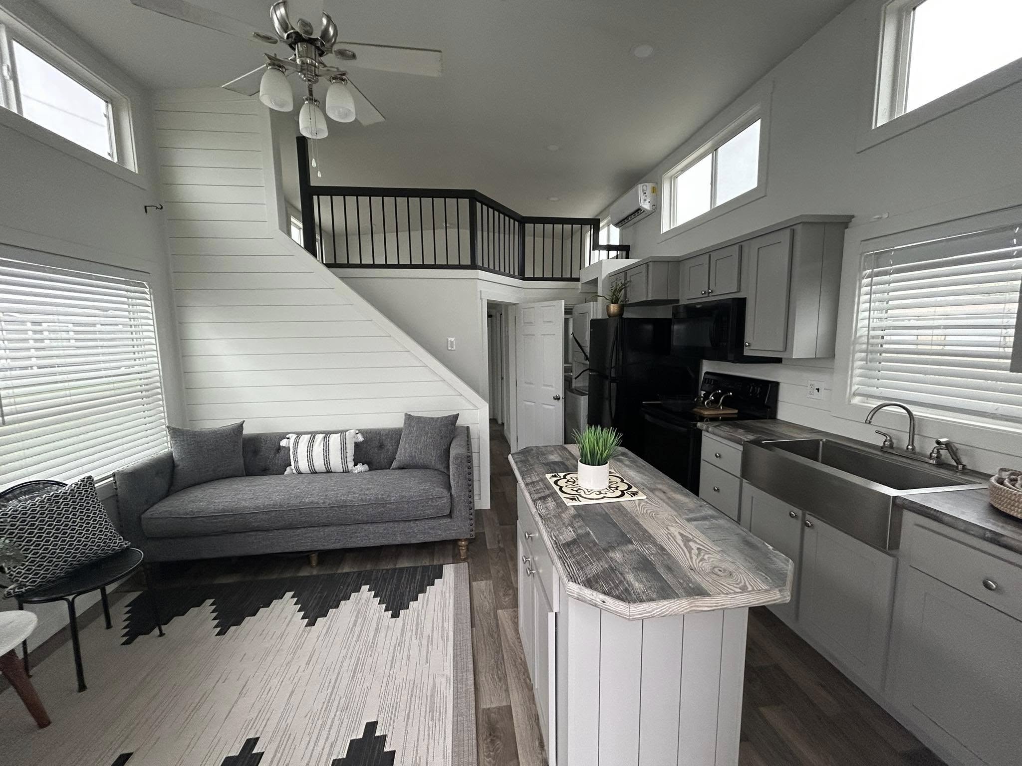 Modern luxury tiny home interior featuring a bright open-concept living area with a grey tufted sofa, a patterned monochromatic area rug, and a rustic kitchen island with a reclaimed wood countertop, showcasing a space-saving loft with black metal safety railings and a sleek grey shaker-style kitchen with a farmhouse sink and stainless steel appliances under a vaulted white shiplap ceiling for a sophisticated move-in ready turn-key residential design.