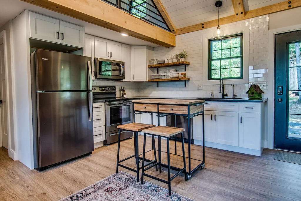Modern kitchen with white cabinets, stainless steel appliances, and wood accents. A small island with stools, wooden beams, and a large window offer a cozy, rustic feel.