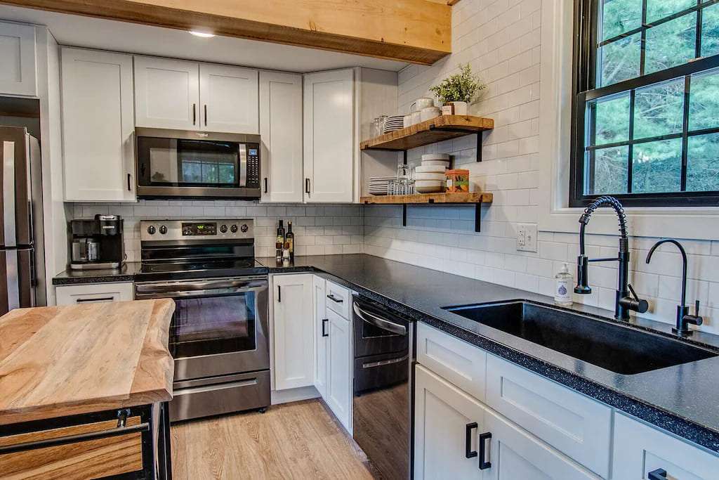 Modern kitchen with stainless steel appliances, white cabinets, and black countertops. Wooden shelves hold dishes; a window adds natural light.