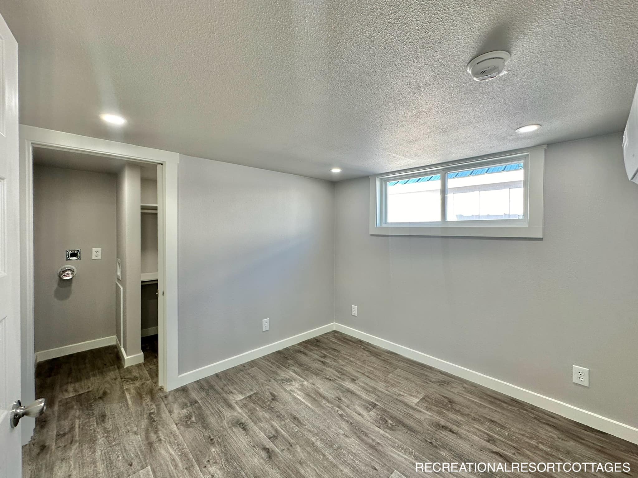 Small, modern room with gray walls and wood-style flooring. Features a high window, recessed lighting, and an open closet. Bright, minimalist feel.