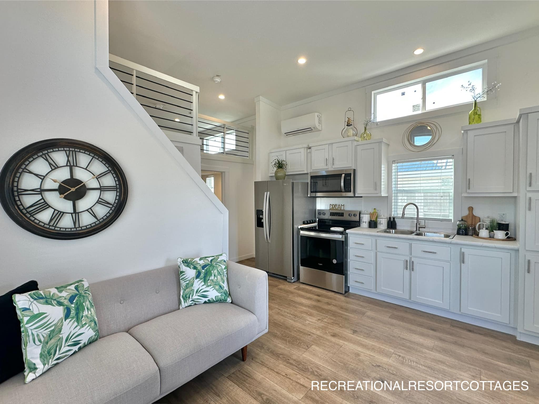 Modern tiny home interior featuring an open kitchen with stainless steel appliances, white cabinets, a cozy sofa with leaf-patterned cushions, and a large wall clock, exuding a bright, airy ambiance.