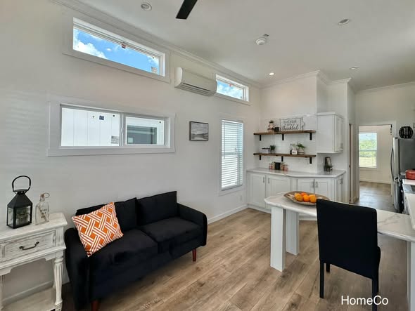 Modern living room and kitchen with natural light. Features a black sofa, orange pillow, white shelves, wood floor, and small dining area with fruit bowl.
