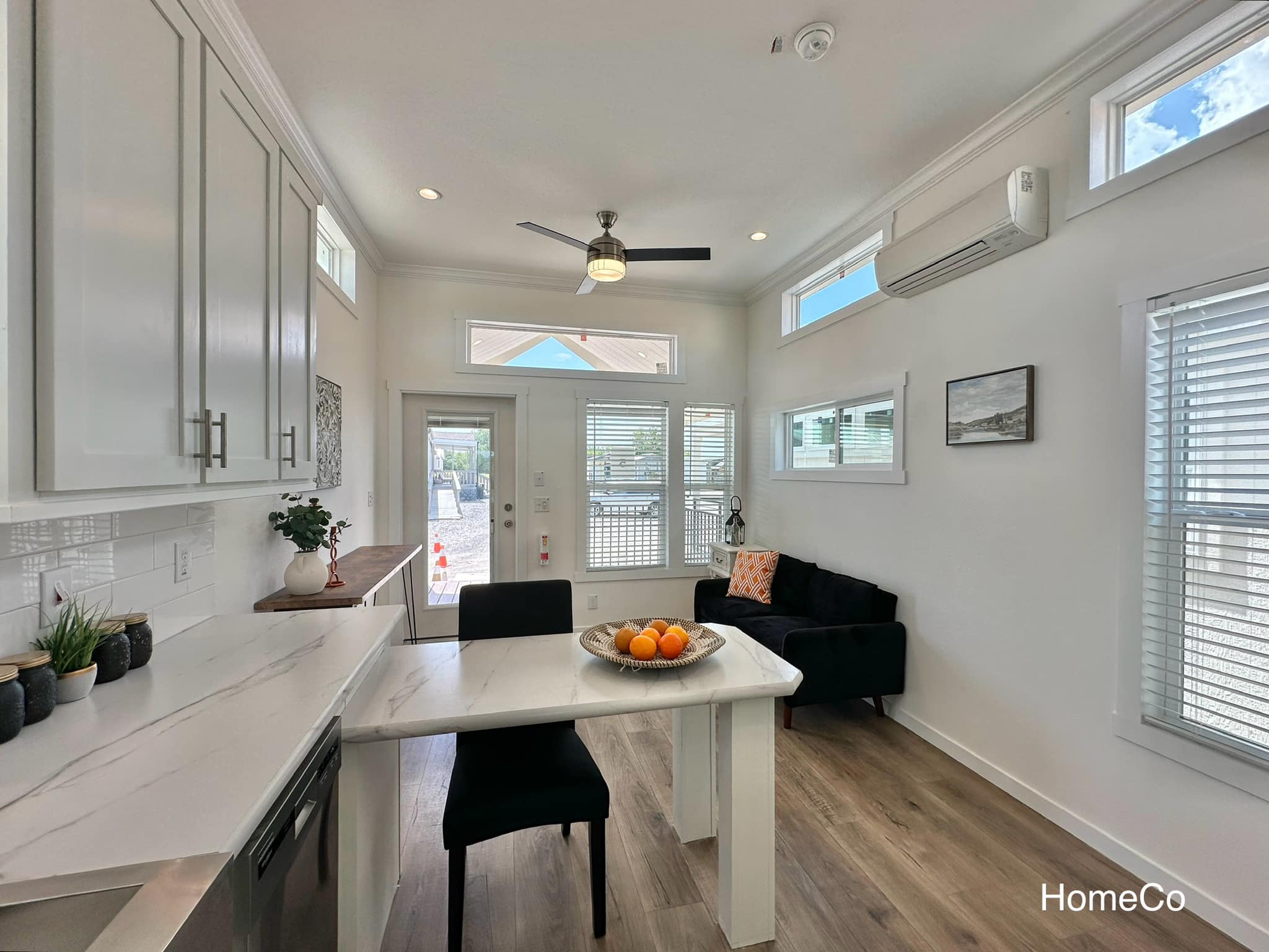 Bright, modern living area with white walls, large windows, ceiling fan, and sleek furnishings. A black sofa and a kitchen island with a bowl of oranges add contrast.