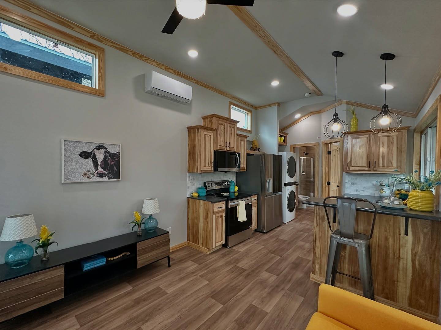 Modern luxury tiny home interior featuring a compact gourmet kitchen with natural wood cabinetry, stainless steel appliances, and a breakfast bar with a metal industrial stool, all set within a bright open-concept living area showcasing a vibrant yellow sofa, wood-grain vinyl flooring, and stylish cage-style pendant lighting under a vaulted ceiling for a functional and sophisticated small-space residential design.