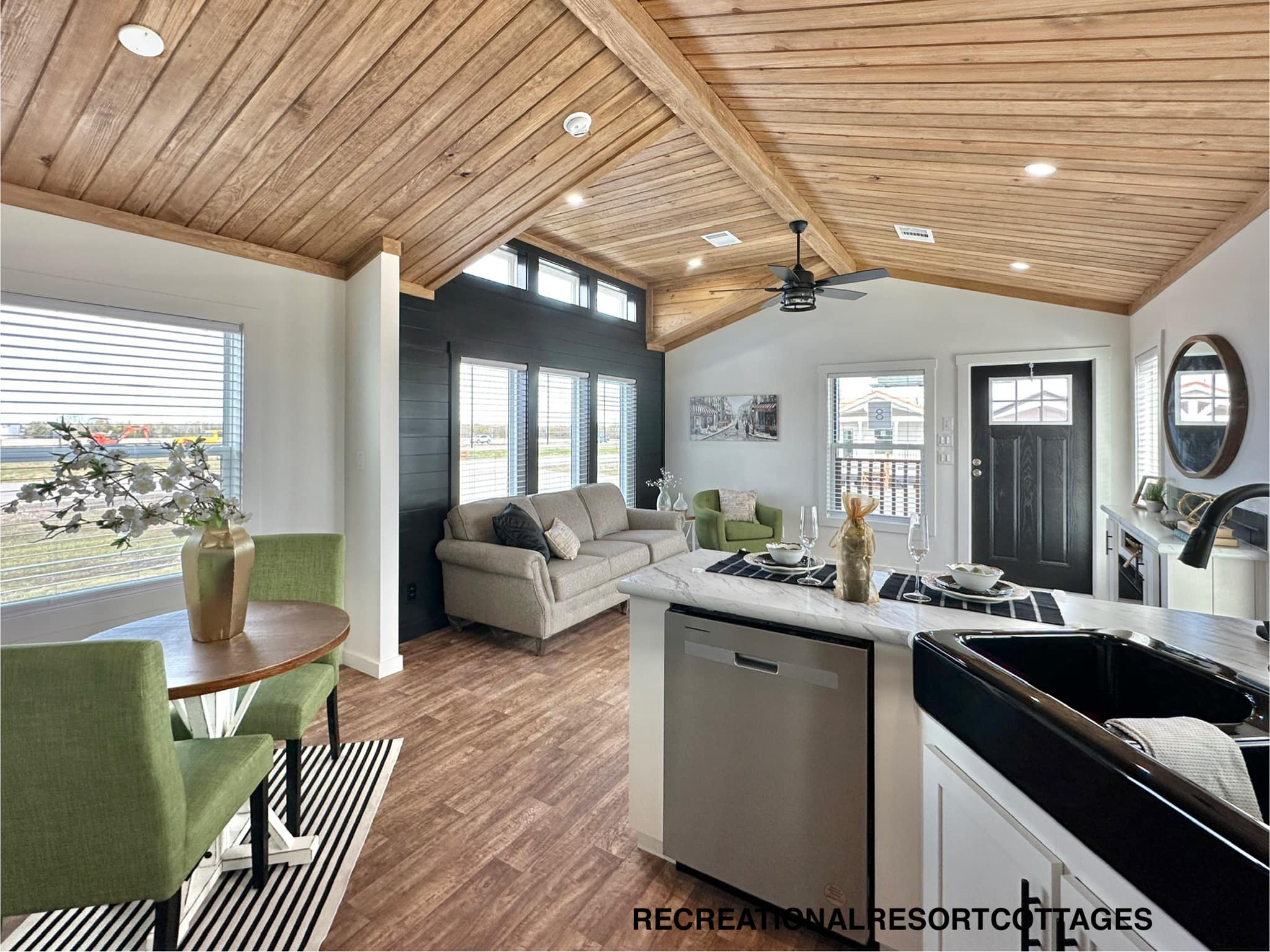 Open-concept living area with vaulted wooden ceiling, featuring a cozy seating area, dining nook with green chairs, and modern kitchen. Bright and inviting.