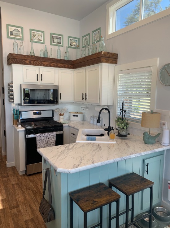 A cozy kitchen features white cabinets, a marbled countertop, and teal base. Wooden stools, decor items, and a window create a warm, inviting atmosphere.