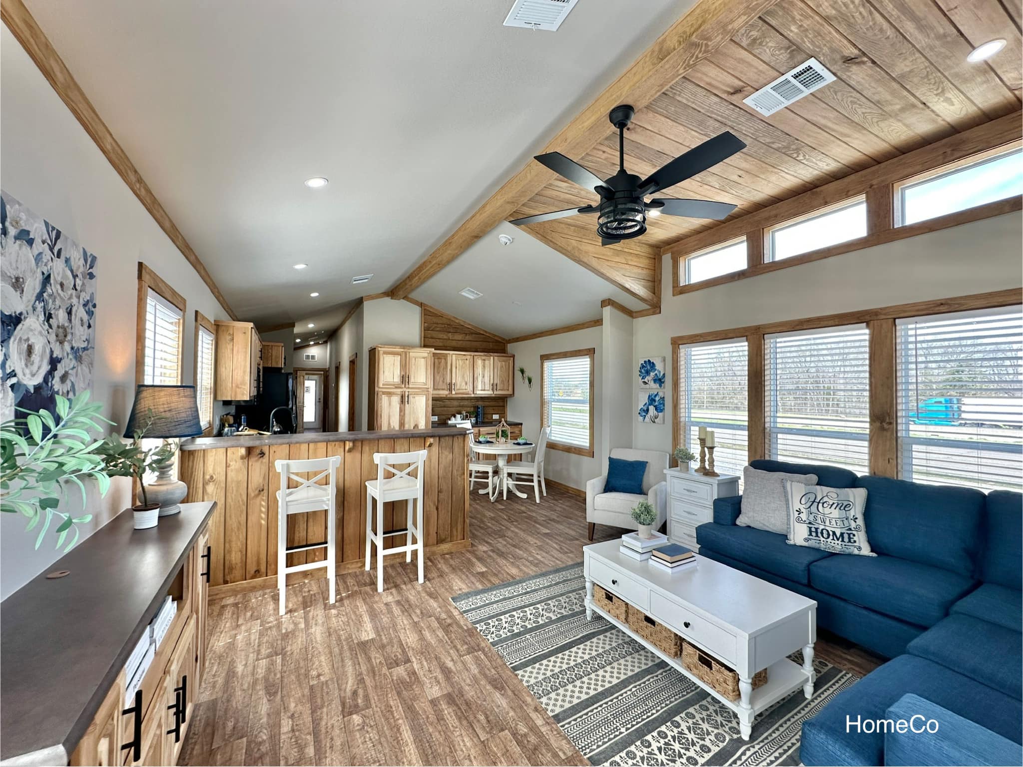 Spacious living area with wood accents, featuring a blue sofa, patterned rug, and white coffee table. Kitchen with bar stools and dining area visible. Cozy, inviting atmosphere.