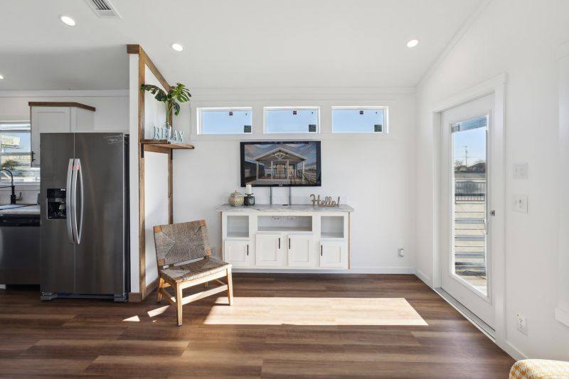Bright, modern interior featuring a stainless steel fridge, wooden chair, and white cabinet beneath a landscape photo. Warm, inviting atmosphere.