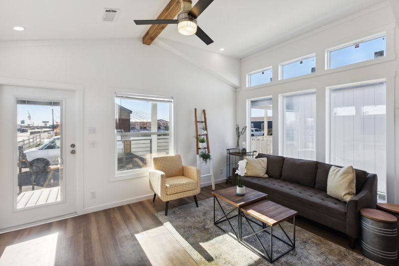 Bright and airy living room in a modern tiny home, featuring a vaulted ceiling with exposed wooden beams and a stylish ceiling fan, a large dark gray sofa with cream pillows, a matching armchair with a checkered pattern, and a rustic wooden ladder shelf, all illuminated by abundant natural light from numerous large windows, creating a welcoming and spacious feel.