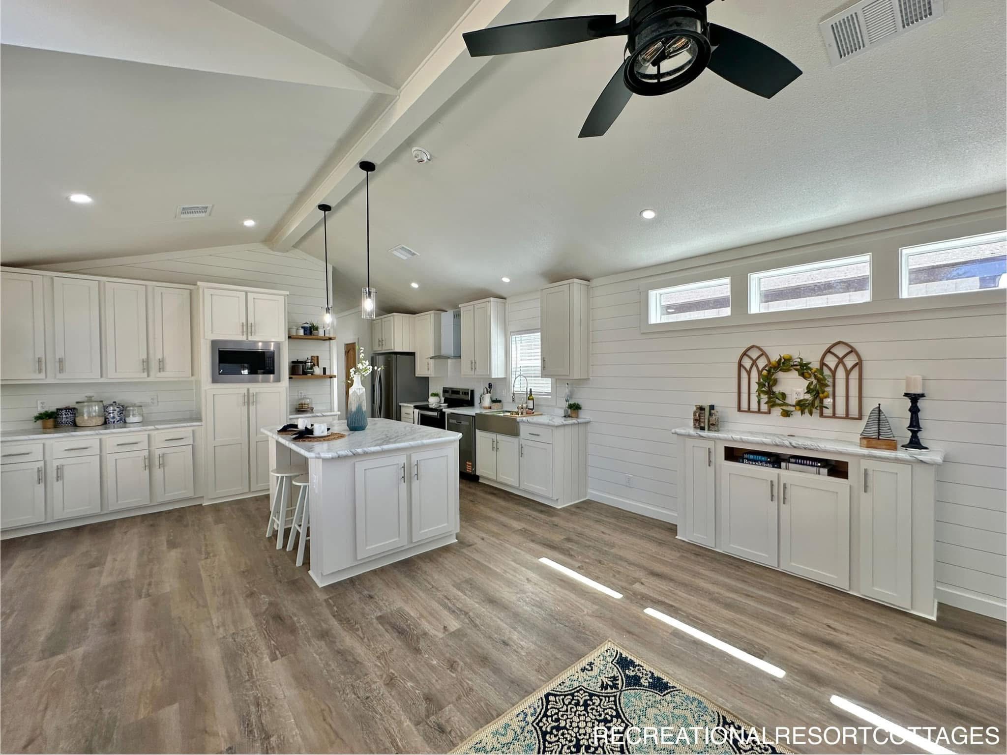 Spacious farmhouse-style kitchen with white cabinetry, center island with stools, stainless steel appliances, and a ceiling fan. Bright and inviting.