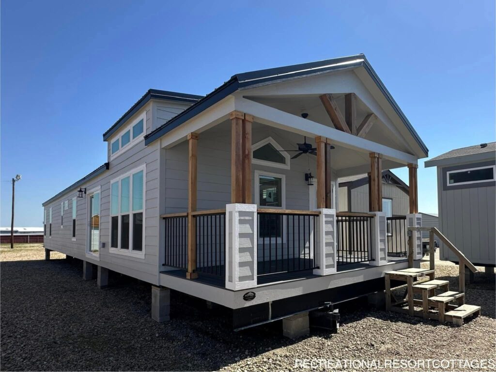 A modern tiny home with light gray siding and a dark metal roof sits elevated on a gravel lot. A covered porch with wooden pillars and black railings adds charm under a clear blue sky.