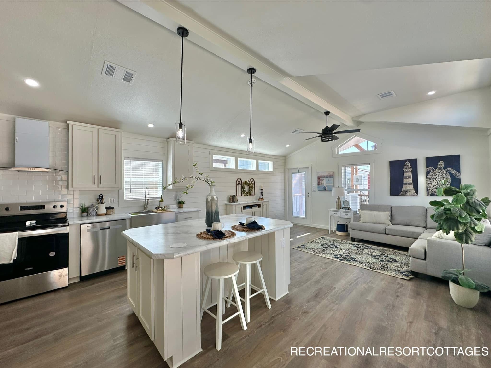 Modern open-plan kitchen and living room with vaulted ceilings. Features white cabinets, island with stools, stainless steel appliances, and cozy gray sofa.